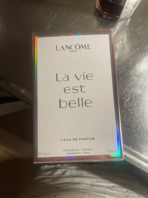 La Vie Est Belle parfym från Lancôme - Elegant parfymflaska från Lancôme, La Vie Est Belle, med rosa doftvätska. Flaskan är fyrkantig med rundade kanter och har ett dekorativt silverfärgat band runt halsen samt en transparent kork. Perfekt för dig som gillar lyxiga och feminina dofter. Helt ny fortfarande i förpackningen och plast över, 100 ml 