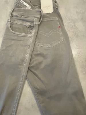 Grå jeans från Replay - Snygga grå jeans från Replay med klassisk femficksdesign och diskreta sömmar. Jeansen har en liten röd detalj på bakfickan och är tillverkade i ett mjukt, stretchigt material för extra komfort. Perfekta för en stilren och avslappnad look. Nu tvättade 
