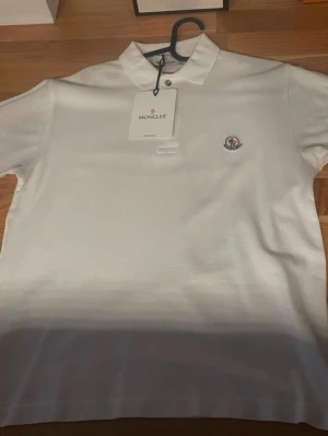 Vit Moncler pikétröja i bomull - Klassisk vit pikétröja från Moncler med liten logga på bröstet och krage med knapp. Tillverkad i mjuk bomull och har korta ärmar. Perfekt för en clean och stilren look.