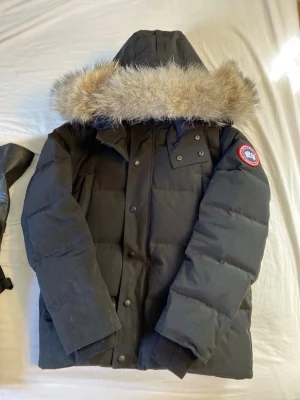 Svart dunjacka från Canada Goose - Svart dunjacka från Canada Goose med huva och avtagbar pälskant. Klassisk patch på ärmen, stora fickor framtill och tryckknappar. Perfekt för kalla vinterdagar och riktigt varm tack vare dunfyllningen.