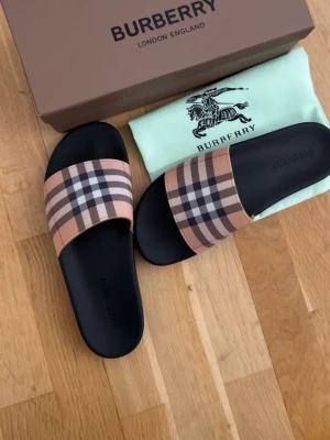 Burberry tofflor  - Snygga sandaler från Burberry med det ikoniska beige, svarta och röda rutmönstret på remmen. Sulan är svart och har en stilren, enkel design. Perfekta för dig som vill ha en lyxig och trendig look till sommaren.