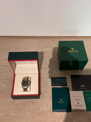 Role Daytona guld och silver klocka - Snygg Role Daytona med svart urtavla och detaljer i både guld och silver. Länken är tvåfärgad i stål och guld, och klockan har flera kronograffunktioner. Kommer med originalbox och certifikat. Perfekt accessoar för dig som gillar exklusiv stil.