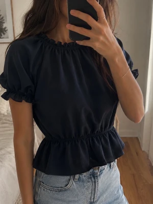 Mörkblå blus med volangdetalj - Snygg mörkblå blus med korta puffärmar och volang i midjan. Blusen har rund halsringning med rynkade detaljer och är perfekt att styla med jeans eller kjol. Materialet känns mjukt och lätt, vilket gör den skön att bära.