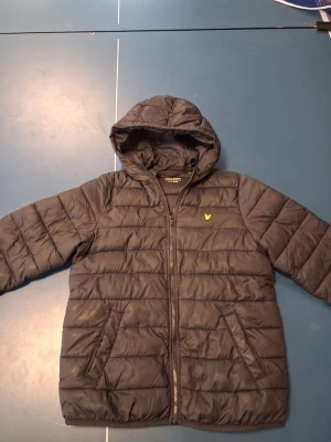 Svart pufferjacka från Lyle & Scott - Snygg svart pufferjacka från Lyle & Scott med huva och dragkedja framtill. Jackan har två fickor och det klassiska gula logotypbroderiet på bröstet. Perfekt för kalla dagar och riktigt skön att bära. Materialet är lätt och värmande.