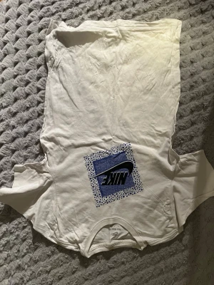 Vit Nike t-shirt med blått tryck - Vit t-shirt från Nike med ett stort blått och svart tryck på bröstet. Trycket har ett rutmönster med prickar och den klassiska Nike-loggan. T-shirten har rund hals och korta ärmar, perfekt för en avslappnad streetstil.