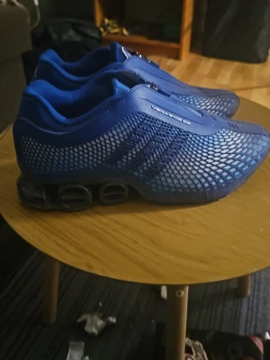 Blå Adidas Megaride sneakers - Snygga blå Adidas Megaride sneakers med futuristisk mesh-design och dragkedja framtill. Skorna har en låg profil och tydliga dämpande sulor med synliga fjädrar baktill. Materialet är en kombination av syntet och mesh för bra andningsförmåga och komfort.