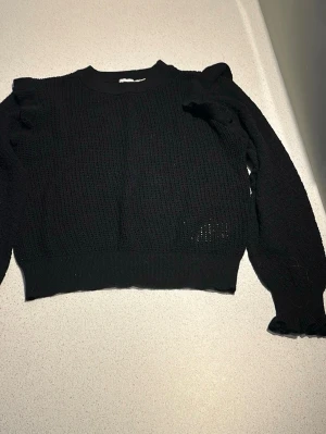 Svart stickad tröja med volang - Svart stickad tröja från Carin Wester med volangdetaljer på axlarna och ribbade muddar. Tröjan har en rund halsringning och är tillverkad i ett mjukt, luftigt material. Perfekt för dig som gillar stilrena och trendiga plagg.