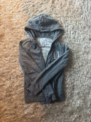 Grå hoodie från Armani Exchange - Snygg grå hoodie från Armani Exchange med dragkedja och huva. Mjuk bomullsblandning och logga 'A|X' på bröstet. Perfekt för en avslappnad och stilren look.