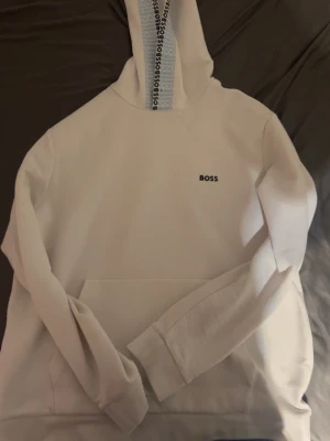 Vit hoodie från BOSS med logga - Snygg vit hoodie från BOSS med liten svart logga på bröstet och BOSS-tryck längs insidan av huvan. Klassisk känguruficka framtill och ribbade muddar. Perfekt för en clean och stilren look.