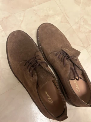 Bruna mockaloafers från Clarks - Snygga bruna loafers från Clarks i mocka med klassisk design och rund tå. Skorna har snörning och en robust sula med vit söm längs kanten. Perfekta för dig som gillar stilrena och tidlösa skor med en modern touch.