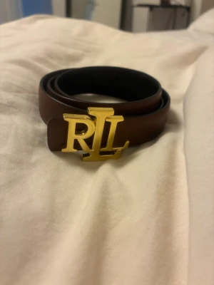 Brunt skärp från Ralph Lauren - Snyggt brunt skärp från Ralph Lauren med ett stort guldfärgat RL-spänne. Skärpet är i slätt skinn och passar perfekt för att lyfta vilken outfit som helst. Klassisk och stilren design med exklusiv känsla.