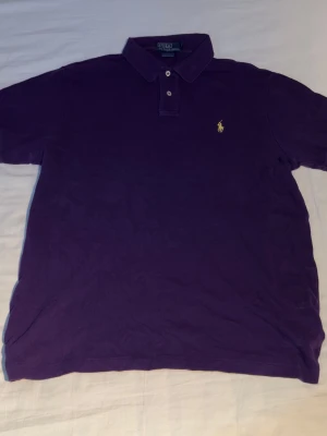 Lila pikétröja från Polo Ralph Lauren - Säljer en klassisk lila pikétröja från Polo Ralph Lauren med gul broderad logga på bröstet. Tröjan har korta ärmar, krage och knappar framtill. Perfekt för en stilren och avslappnad look.