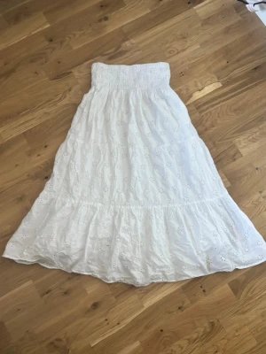 Vit långkjol med broderade detaljer - Luftig vit långkjol med smock i midjan och broderade hålmönster över hela kjolen. Kjolen har volang längst ner och är perfekt för varma dagar. Tillverkad i bomull för en skön och sval känsla.