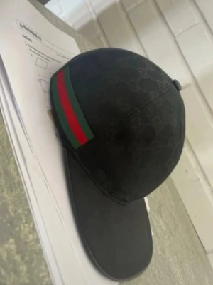 Svart Gucci keps med GG-mönster - Snygg svart keps från Gucci med klassiskt GG-mönster över hela kepsen och en grön-röd rand på sidan. Kepsen har böjd skärm och är tillverkad i ett tygmaterial som känns lyxigt och trendigt. Perfekt accessoar för att lyfta din streetwear-stil.