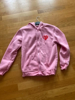 Rosa hoodie med hjärtdetalj - Säljer en rosa hoodie från KappAhl med dragkedja och fickor framtill. På bröstet finns ett rött hjärta med ögon som ger en unik och lekfull touch. Hoodien har huva med dragsko och är tillverkad i mjukt sweatshirtmaterial. Perfekt för en avslappnad och trendig stil.