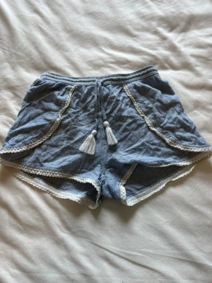Blå shorts med spets och tofsar - Supersöta blå shorts med vita spetsdetaljer längs kanterna och två vita tofsar framtill. Shortsen har en avslappnad passform och är perfekta för varma dagar. Materialet känns mjukt och luftigt, och de har en bohemisk vibe. Passar folk som bär XSS/XS