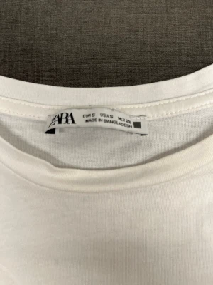 Vit t-shirt med spetsdetalj från Zara S - Vit t-shirt från Zara i mjuk bomull med korta ärmar. Ärmsluten har en snygg svart spetskant som ger en unik touch. Enkel och stilren modell som passar till det mesta. Perfekt för dig som gillar klassiska plagg med en twist. Storlek S. 