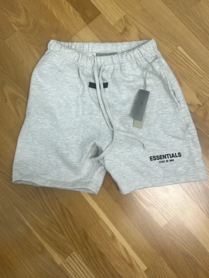 Grå Essentials shorts med snörning - Snygga ljusgrå shorts från Fear of God Essentials med svart logga på benet. Shortsen har elastisk midja med snörning och sidofickor. Perfekta för en avslappnad och trendig stil.