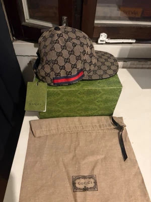 Gucci GG Supreme keps beige - Säljer en beige Gucci keps i GG Supreme-mönster med blå och röda ränder på sidan. Kepsen har böjd skärm och justerbar rem bak. Tillverkad i canvasmaterial och levereras med originalpåse och box. Perfekt för dig som vill ha en lyxig och trendig accessoar.