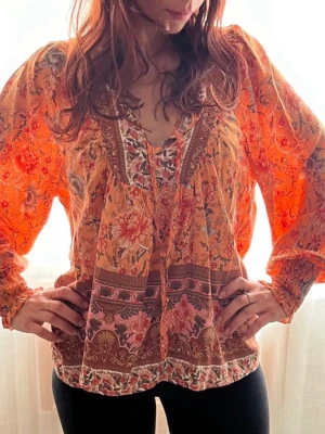 Bohemisk blommig blus i orange toner - Bohemisk blus i tunnt, flowy material med orange botten och blommigt mönster i rosa, rött och brunt. V-ringad med knytband och långa, puffiga ärmar med smock vid ärmslut. Perfekt för dig som gillar färg och mönster.