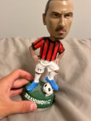 Fotbollsspelare figur med boll - Säljer en cool figur av en fotbollsspelare i röd och svart randig tröja, vita shorts och blå skor. Figuren står på en grön platta med text och har en fot på en fotboll. Perfekt för dig som gillar fotboll och samlarprylar.