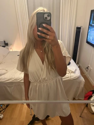 beige strandklänning  - Säljer en beige strandklänning med omlottdesign och resår i midjan. Klänningen har korta ärmar, v-ringning och öppen rygg med knytband i nacken. Perfekt för sommarens varma dagar och enkel att styla med accessoarer.