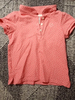Rosa prickig pikétopp med hjärtdetalj - Söt rosa pikétopp med vita prickar och liten broderad hjärtdetalj på bröstet. Toppen har korta ärmar, krage och knappar framtill. Perfekt för dig som gillar en lekfull och avslappnad stil.