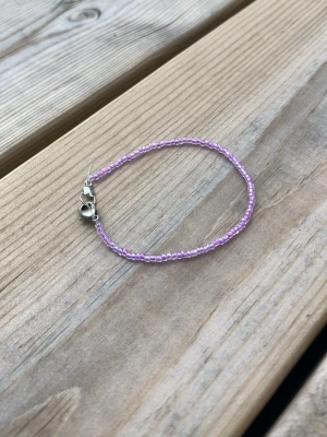 Lila pärlarmband med silverlås - Ett tunt armband med små lila pärlor och ett silverfärgat hjärtformat lås. Perfekt för dig som gillar enkla och söta accessoarer. Armbandet har en minimalistisk design och passar till många olika stilar.