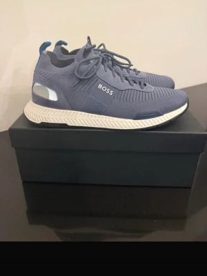 BOSS Skor - Snygga blå sneakers från BOSS med stickad ovandel och vita sulor. Skorna har reflekterande detaljer på hälen, blå hälflik och tydlig BOSS-logga på sidan och baksidan. Perfekta för dig som gillar stilrena och moderna sneakers.