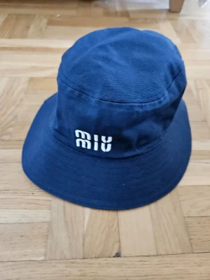 Mörkblå bucket hat från Miu Miu - Snygg mörkblå fiskar hatt från Miu Miu med broderad vit logga framtill. Hatten är tillverkad i ett kraftigt tyg och har en klassisk form med bred brätte. Perfekt accessoar för att lyfta din stil.