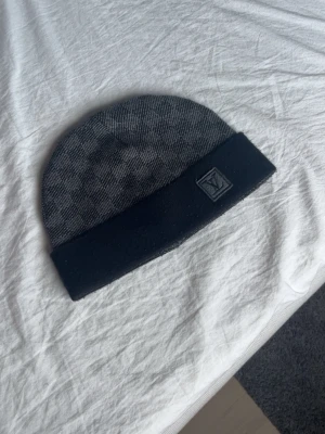 Grå mössa - Säljer en snygg grå mössa med diskret rutmönster och svart uppvikt kant. Mössan har en klassisk patch med logga framtill och är tillverkad i mjuk ull som håller dig varm under kyliga dagar. Obs ej äkta används få par gånger