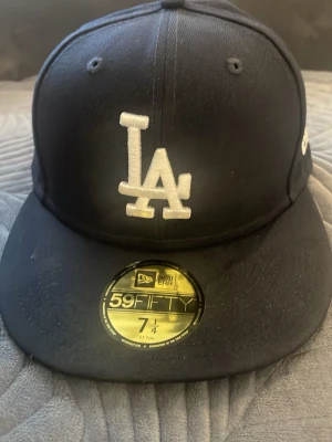 Svart LA-keps från New Era 59FIFTY - Säljer en svart keps från New Era 59FIFTY med LA-logga broderad i vitt framtill. Klassisk flat brim och broderad New Era-logga på sidan. Tillverkad i ett slitstarkt tyg, perfekt för dig som gillar streetwear och sportig stil.