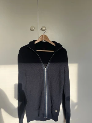 Mörkblå hoodie med dragkedja - Snygg mörkblå hoodie med dragkedja framtill och huva. Tillverkad i ett stickat material som ger en avslappnad och trendig look. Perfekt för lager på lager och passar till det mesta i garderoben. Långa ärmar och diskret detalj vid nederkanten.