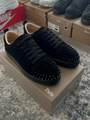 Svarta Louboutin sneakers  - Säljer ett par svarta sneakers från Christian Louboutin med mattsvarta nitar över tåpartiet och sidorna. Skorna är låga, har svarta skosnören och är tillverkade i mocka med läderdetaljer. Insidan är beige och sulan är svart. Riktigt edgy och unika sneakers för dig som vill sticka ut.