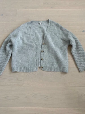 Ljusgrå stickad kofta från GAP - Ljusgrå stickad kofta från GAP med ribbade muddar och knappar framtill. Koftan har en avslappnad passform, v-ringning och är tillverkad i ett mjukt garn som känns skönt mot huden. Perfekt att slänga över en t-shirt eller topp.
