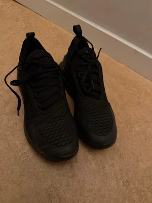 Svarta Nike Air Max 270 sneakers - Säljer ett par helsvarta Nike Air Max 270 sneakers med mesh-ovandel och synlig Air-enhet i hälen. Skorna har en sportig look med snörning och dragflik bak. Perfekta för dig som gillar streetwear och vill ha bekväma, stilrena sneakers.