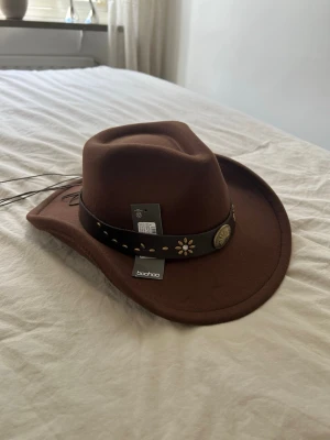 Brun cowboyhatt från Boohoo - Säljer en brun cowboyhatt från Boohoo med dekorativt band runt hatten, prydd med blommor och metallknapp. Hatten har bred brätte och snörning baktill. Perfekt accessoar för dig som vill sticka ut med westernstil.