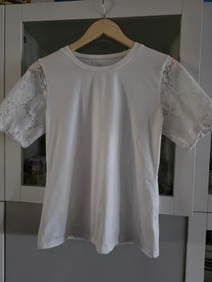 Vit t-shirt med spetsärmar - Snygg vit t-shirt med korta ärmar i spets som ger en romantisk vibe. Kroppen är i mjuk bomull och ärmarna har ett blommigt spetsmönster som sticker ut. Perfekt för dig som vill ha en basic tee med det lilla extra. 