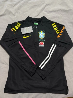 Svart Brasil Nike träningströja - Svart långärmad träningströja från Nike med Brasilien CBF-logga, gula och vita detaljer samt tryck på ärmarna. Halvlång dragkedja framtill och meshmaterial för bra ventilation. Perfekt för fotbollsträning eller sportiga aktiviteter.