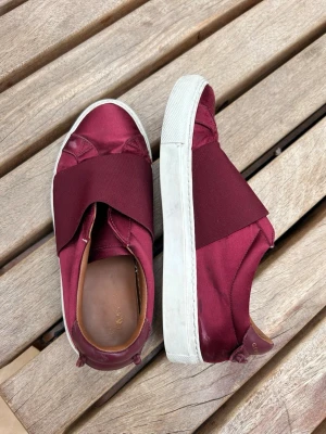 Vinröda Givenchy slip-on sneakers - Snygga vinröda slip-on sneakers från Givenchy med bred resår över foten och vit tjock sula. Skorna har detaljer i läder och textil, samt Givenchy-logga i guld på hälen. Perfekta för dig som vill ha en clean och lyxig look.