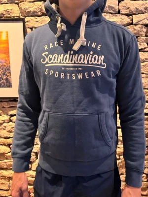 Blå hoodie race marine  - Mörkblå hoodie från Scandinavian Sportswear med vit text på bröstet och stora snören i huvan. Klassisk känguruficka framtill och ribbade muddar vid ärmar och midja. Perfekt för en avslappnad och sportig stil.