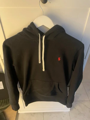 Svart hoodie från Polo Ralph Lauren - Säljer en svart hoodie från Polo Ralph Lauren med klassisk röd logga broderad på bröstet. Hoodien har vit dragsko, stor känguruficka framtill och mjukt material som känns skönt mot huden. Perfekt för en chill och stilren look.