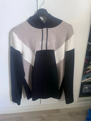 Hugo Boss hoodie i grått och svart - Snygg hoodie från Hugo Boss med grå, svart och vit färgblockdesign. Stor logga på bröstet och justerbar huva med snören. Tillverkad i mjukt material som känns skönt mot huden. Perfekt för en avslappnad och trendig stil.