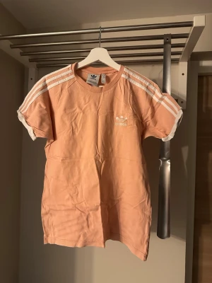 Adidas rosa t-shirt med vita ränder - Snygg rosa t-shirt från Adidas med klassiska vita ränder på axlarna och ärmarna. T-shirten har rund halsringning, korta ärmar och Adidas-logga på bröstet. Perfekt för en sportig och avslappnad stil.