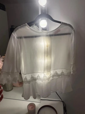 Vit blus med spetsdetaljer - Söt vit blus i transparent material med trekvartsärm och fina spetsdetaljer vid ärmslut och midja. Blusen har rund halsringning och lätt utsvängd nederkant för en romantisk look. Perfekt att styla med jeans eller kjol.