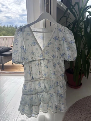 Vit blommig kortklänning med spets - Söt vit kortklänning med blå blommor och puffärmar. Klänningen har v-ringning, spetsdetaljer och volanger i kjolen. Perfekt för sommarens alla tillfällen och ger en romantisk vibe.