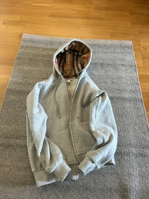 Grå hoodie från Burberry med dragkedja - Snygg grå hoodie från Burberry med dragkedja och klassiskt rutigt foder i huvan. Tröjan har ribbade muddar, dragsko i huvan och två fickor framtill. Perfekt för dig som gillar stilrena och exklusiva plagg med en twist.