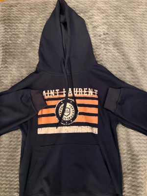 Mörkblå Saint Laurent hoodie - Snygg mörkblå hoodie från Saint Laurent med stor huva och dragsko. Framsidan har ett tryck med orangea och vita ränder samt ett runt emblem i mitten. Hoodien har magficka och ribbade muddar vid ärmar och nederkant.