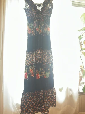 Blommig långklänning med spetsdetaljer - Långklänning med svart bas och blommigt mönster i rosa, grönt och rött. Klänningen har smock upptill, spetsdetaljer vid axelbanden och flera volanger längs kjolen. Den här lite flamenco/marknadsvibe men köpte den i Sverige.💃🏼Knappt använd då jag inte gillar urringningen. Perfekt för en dag på stranden eller en middag ute.🌹