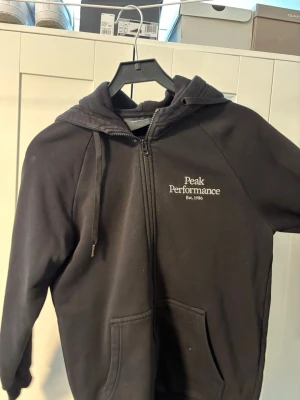 Svart hoodie Peak Performance - Säljer en svart hoodie från Peak Performance med dragkedja framtill och vit logga på bröstet. Hoodien har huva med dragsko och två stora fickor framtill. Perfekt för en avslappnad och sportig stil.